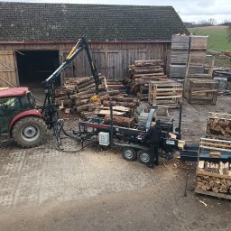 Forest Energy Damian Wantoch-Rekowski - Cięcie drewna na składzie: traktor z dźwigiem podaje kłody do maszyny tnącej, obok skrzynie z pociętym drewnem. W tle budynek gospodarczy i pole.
