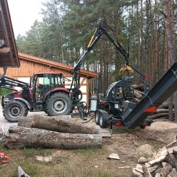 Forest Energy Damian Wantoch-Rekowski - Czerwony traktor z podnośnikiem i przyczepą do cięcia drewna na tle lasu. Na pierwszym planie leżą pnie drzew, obok piła łańcuchowa i stos pociętego drewna.