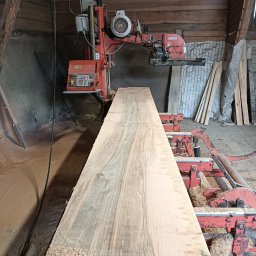 Forest Energy Damian Wantoch-Rekowski - Deska z jasnego drewna na czerwonej maszynie w tartaku. Widoczne trociny i drewniane ściany budynku. Perspektywa z dołu podkreśla długość deski.