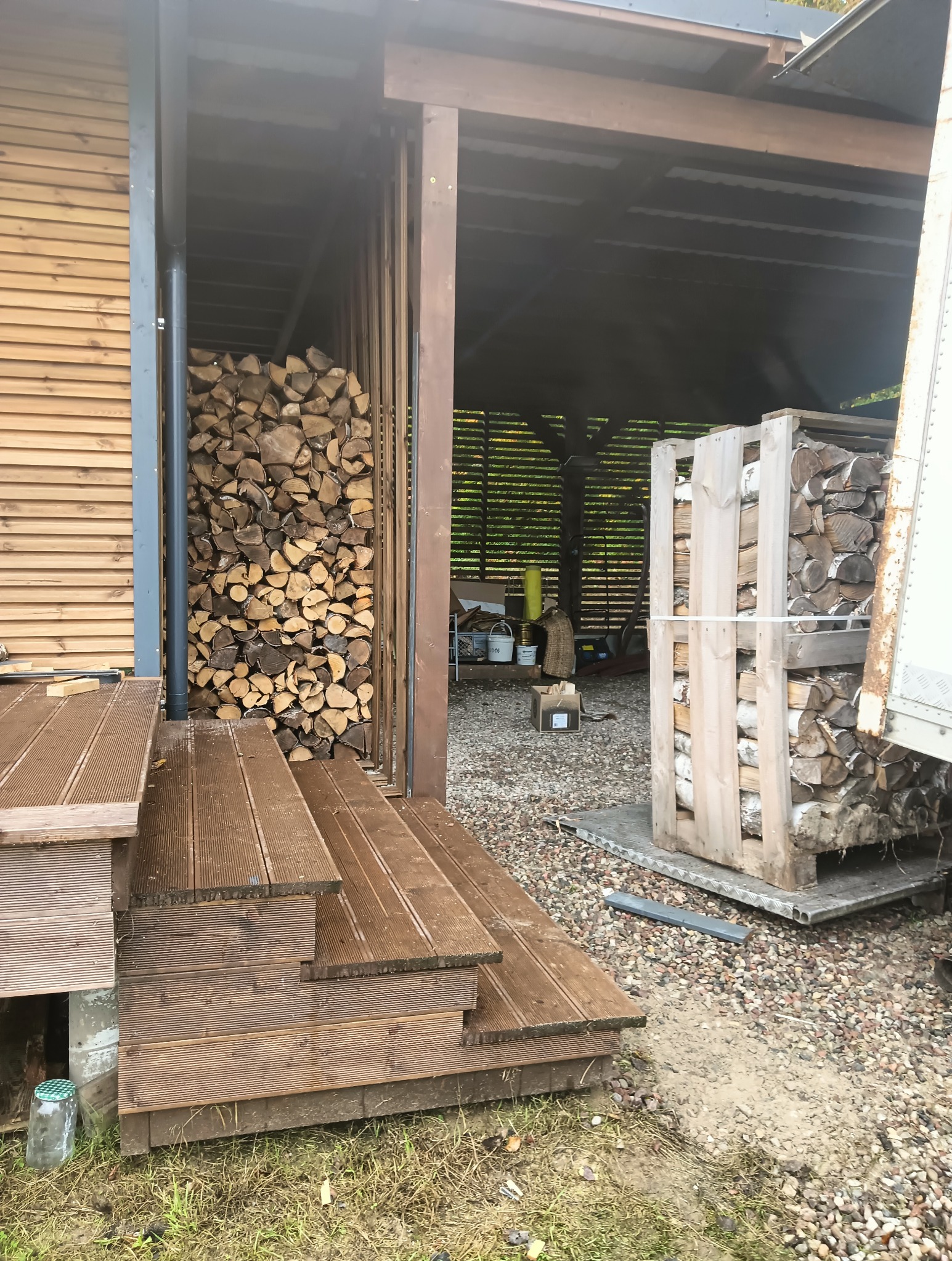 Składowane drewno opałowe w drewnianej konstrukcji, widoczne z zewnątrz przez drewniane schody i taras. Część drewna w palecie. Tło stanowi zadaszona wiata.