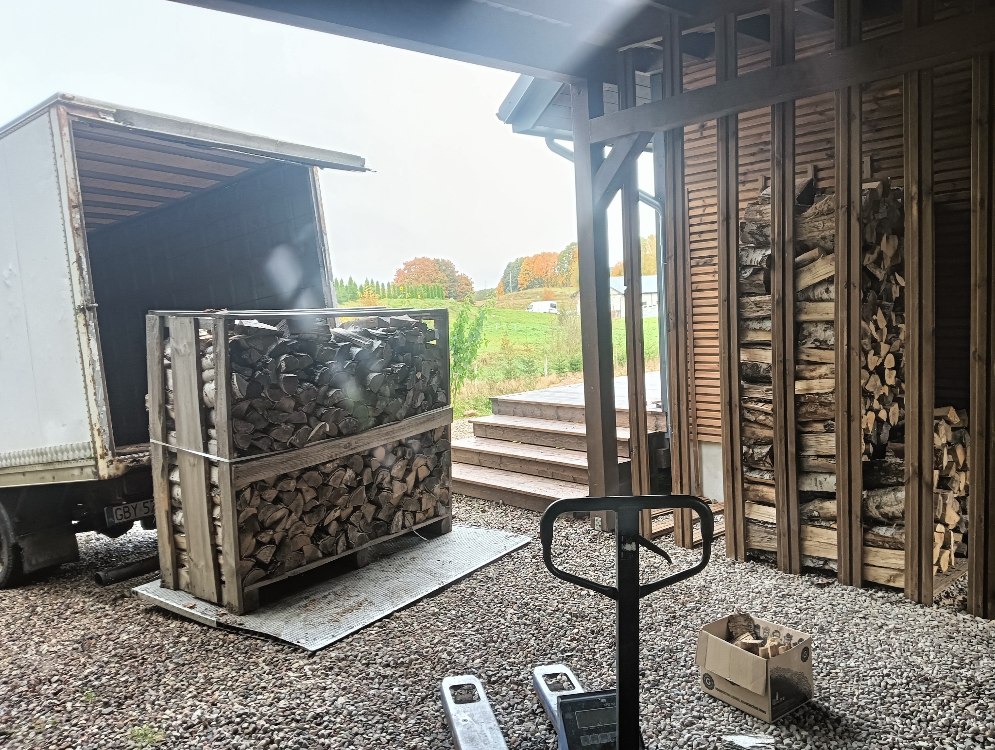 Dostawa drewna opałowego: skrzynia z pociętymi kawałkami drewna na palecie, wyładowywana z ciężarówki na posesję z widokiem na jesienny krajobraz.