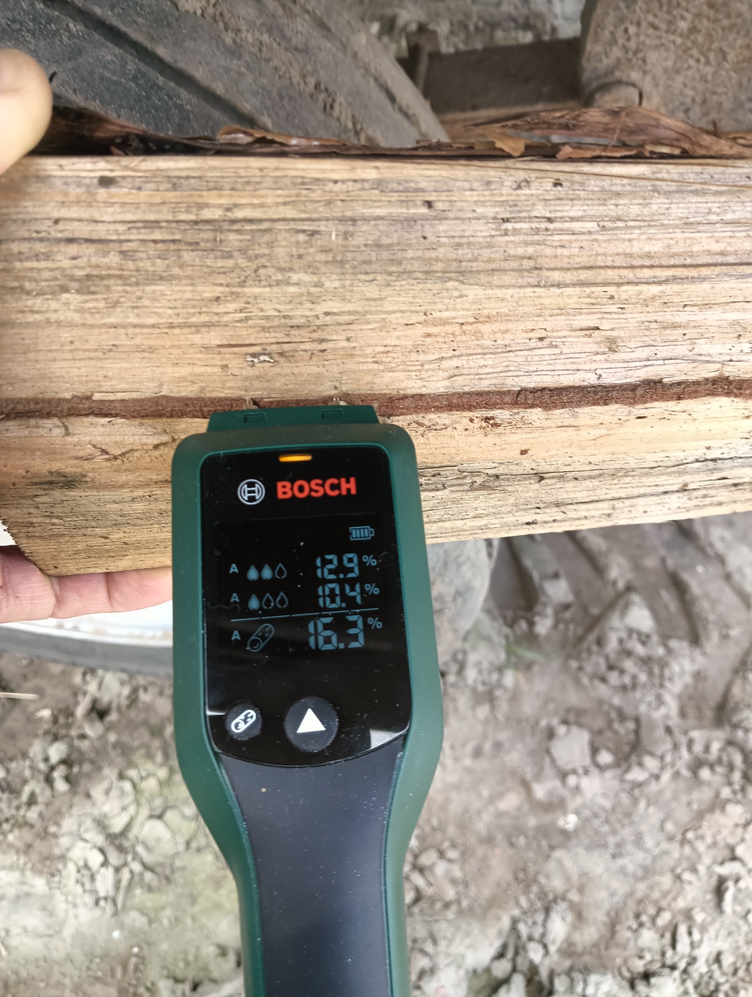 Pomiar wilgotności drewna kominkowego miernikiem Bosch. Wyświetlacz pokazuje 12.9%, 10.4% i 16.3%. Widoczne fragmenty drewna i dłoni. Tło ziemiste.