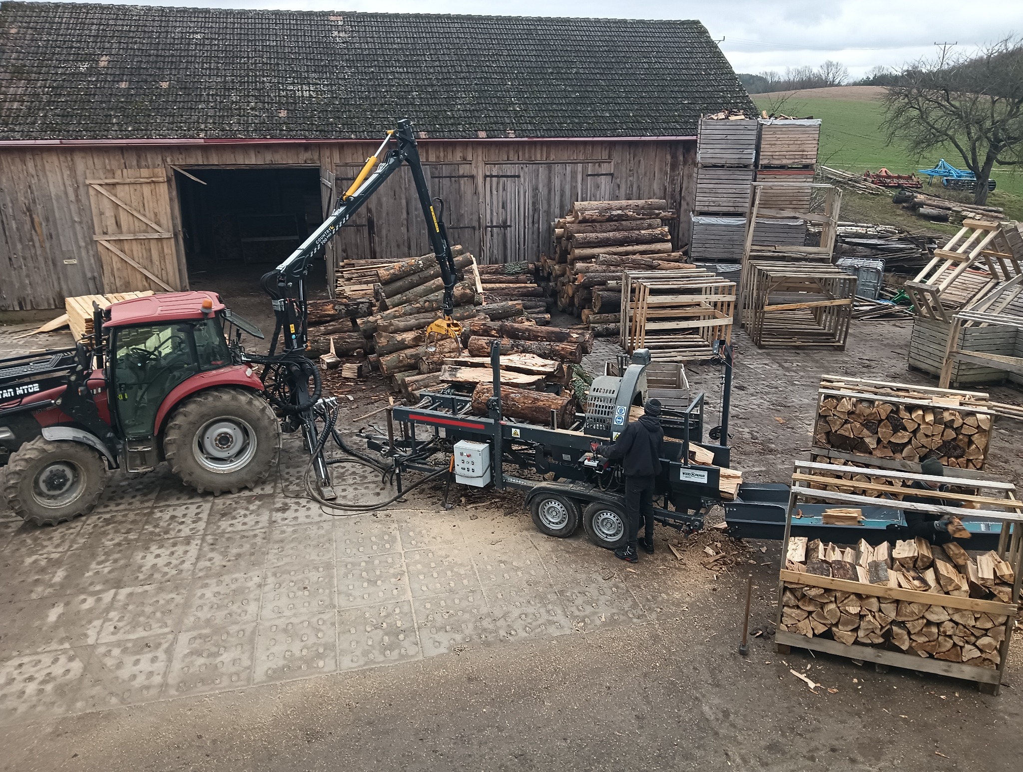Cięcie drewna na składzie: traktor z dźwigiem podaje kłody do maszyny tnącej, obok skrzynie z pociętym drewnem. W tle budynek gospodarczy i pole.