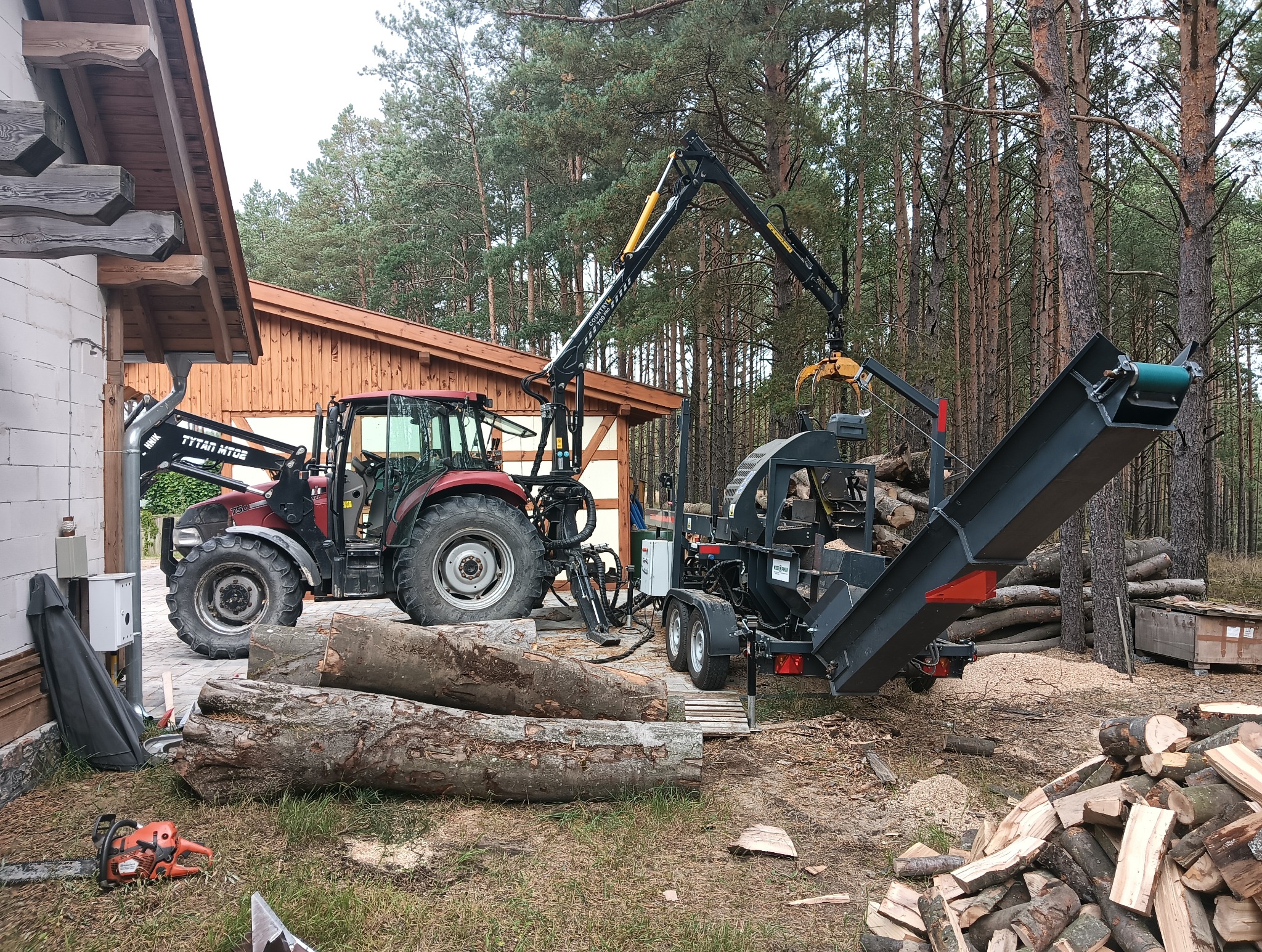 Czerwony traktor z podnośnikiem i przyczepą do cięcia drewna na tle lasu. Na pierwszym planie leżą pnie drzew, obok piła łańcuchowa i stos pociętego drewna.
