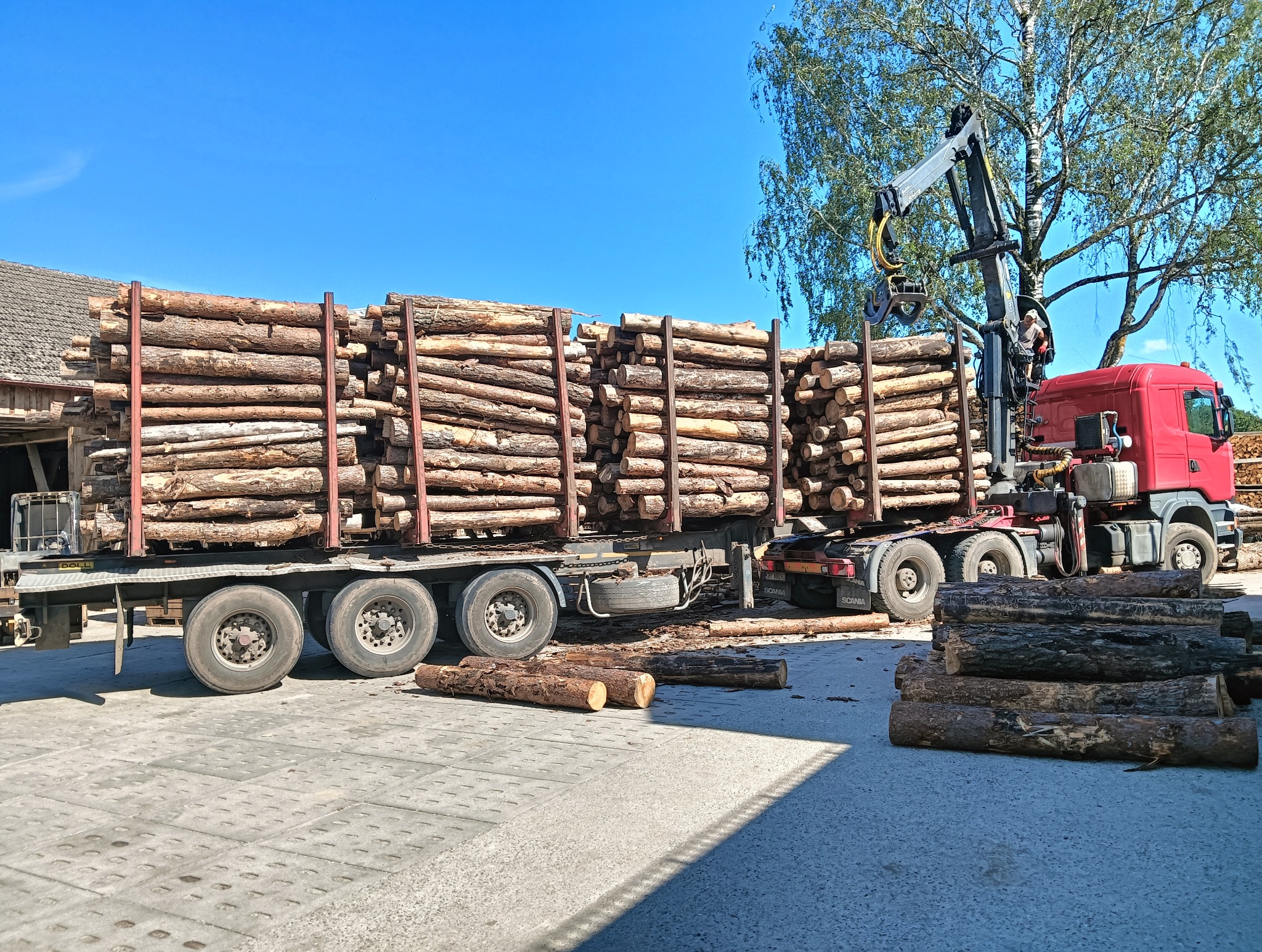Ciężarówka z czerwonym żurawiem załadowana pociętymi kłodami drewna na placu. Na pierwszym planie stos drewna. Błękitne niebo w tle.