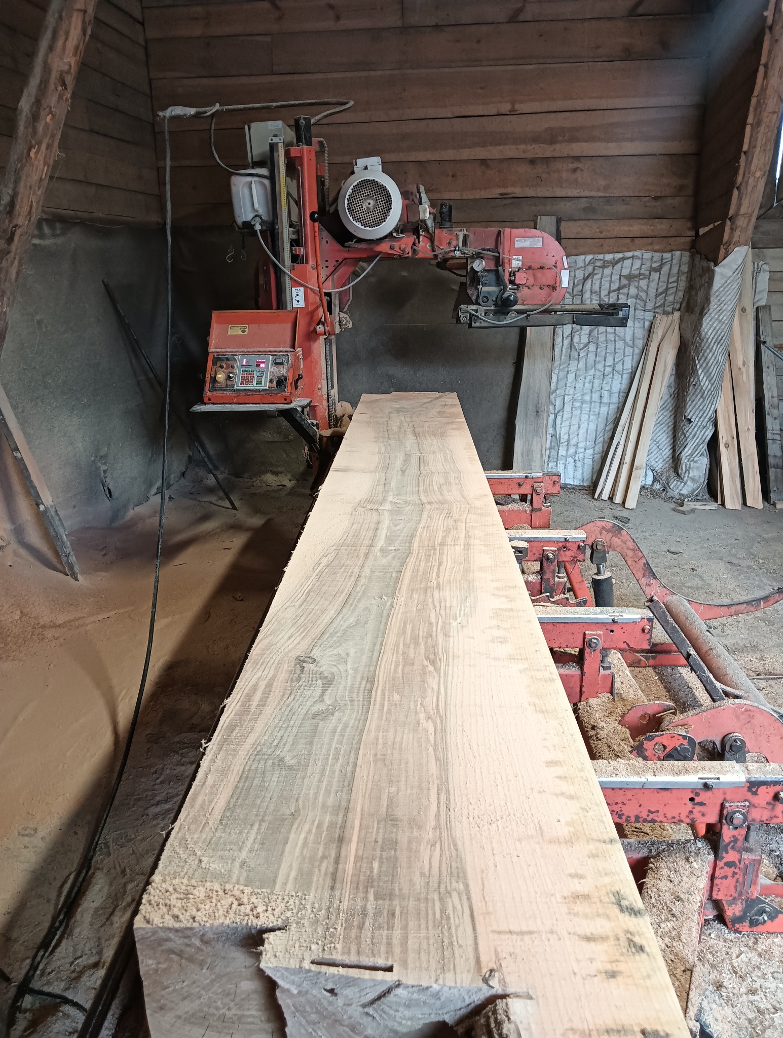 Deska z jasnego drewna na czerwonej maszynie w tartaku. Widoczne trociny i drewniane ściany budynku. Perspektywa z dołu podkreśla długość deski.