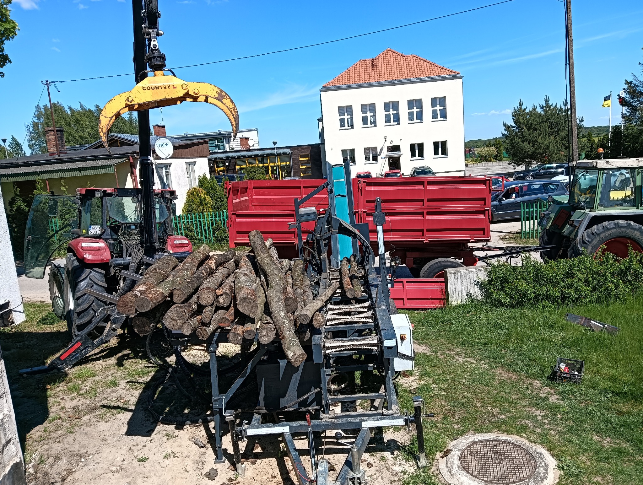 Czerwony traktor z żółtym chwytakiem podnosi kłody drewna na podajnik piły mechanicznej. W tle budynek i przyczepa. Słoneczny dzień.