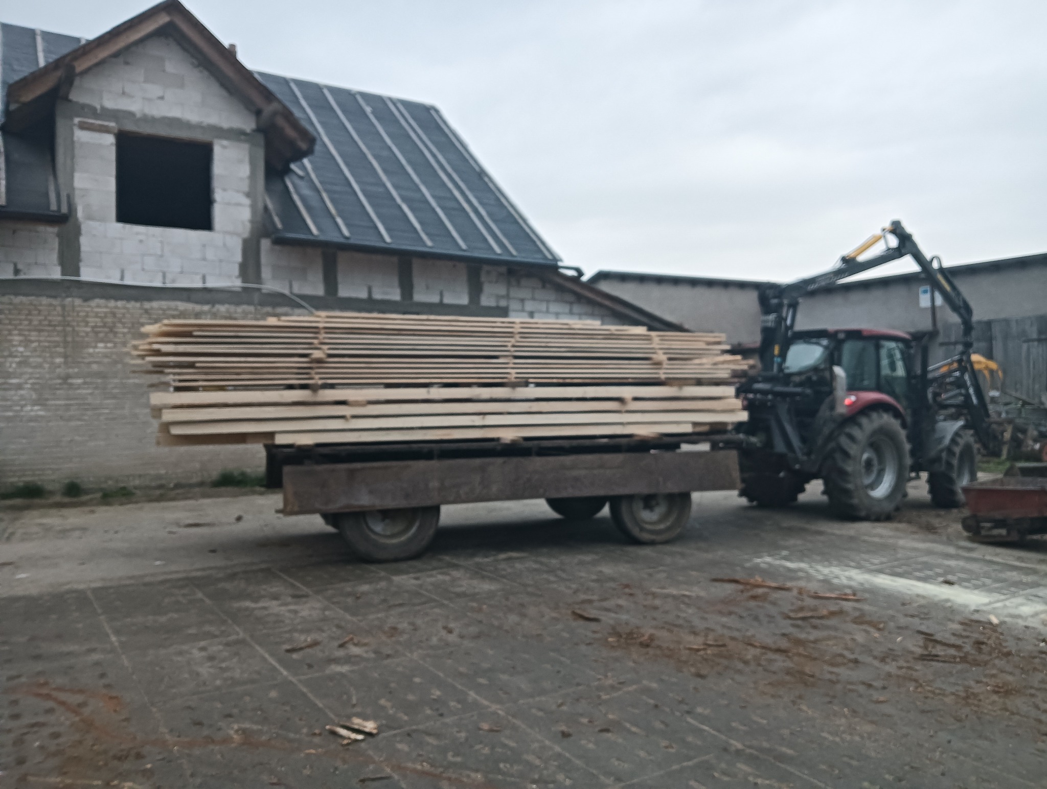 Transport tarcicy traktorem z HDS na tle budynku w trakcie budowy. Drewno ułożone na przyczepie. Pochmurne niebo.
