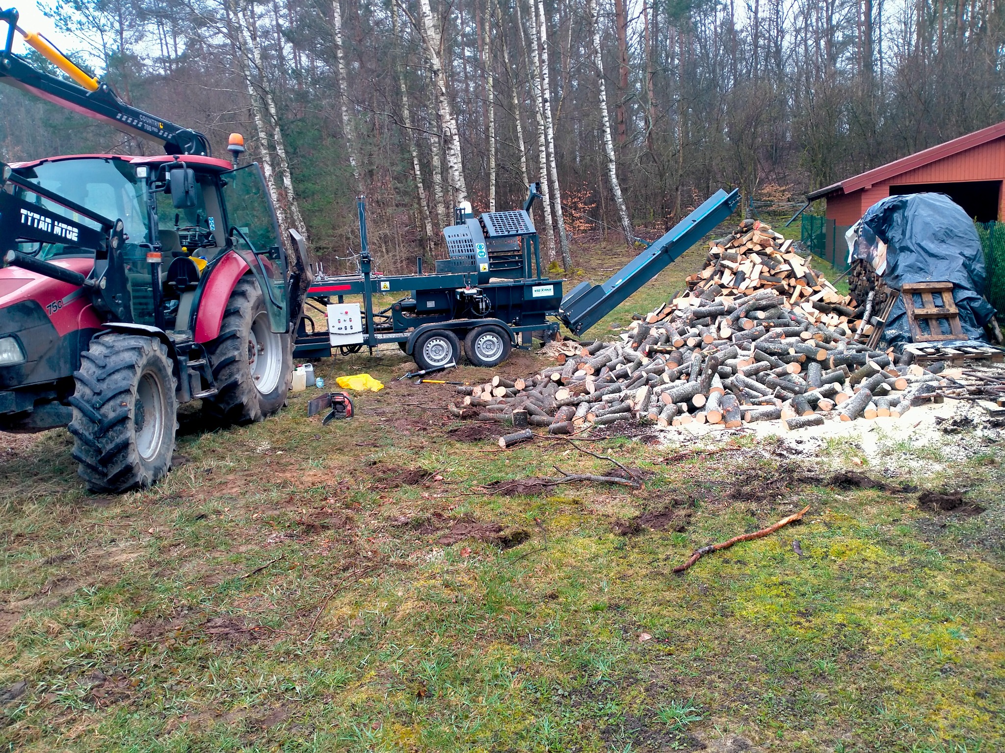 Czerwony traktor z podnośnikiem i przyczepą do cięcia drewna obok sterty pociętych klocków na tle drzew i budynku. Widoczne ślady opon na trawie.