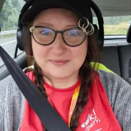 Autoportret kobiety w okularach, czapce i słuchawkach, siedzącej w samochodzie z zapiętym pasem. Widoczna koszulka z logo 'K! tours'.