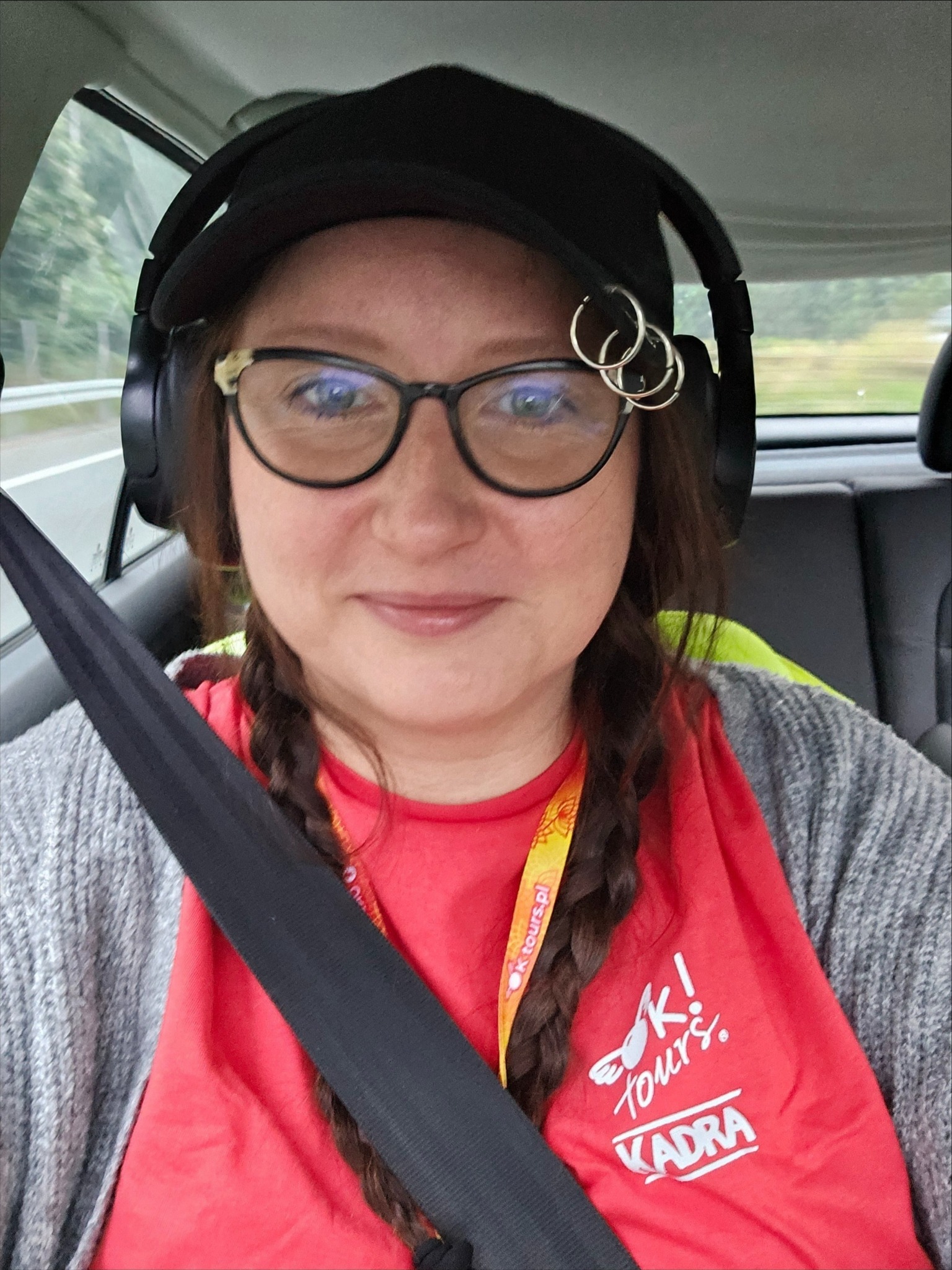 Autoportret kobiety w okularach, czapce i słuchawkach, siedzącej w samochodzie z zapiętym pasem. Widoczna koszulka z logo 'K! tours'.