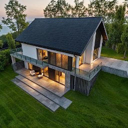 JS Pracownia architektoniczno-budowlana Joanna Syrek - Nowoczesny dom z płaskim dachem i dużymi przeszkleniami, otoczony zielonym trawnikiem i drzewami, widziany z lotu ptaka o zmierzchu. Minimalistyczny design.