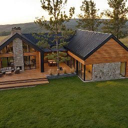 JS Pracownia architektoniczno-budowlana Joanna Syrek - Nowoczesny dom z drewnianą elewacją i kamiennymi elementami, duży taras z meblami wypoczynkowymi, otoczony zielonym trawnikiem i drzewami, widok z góry.