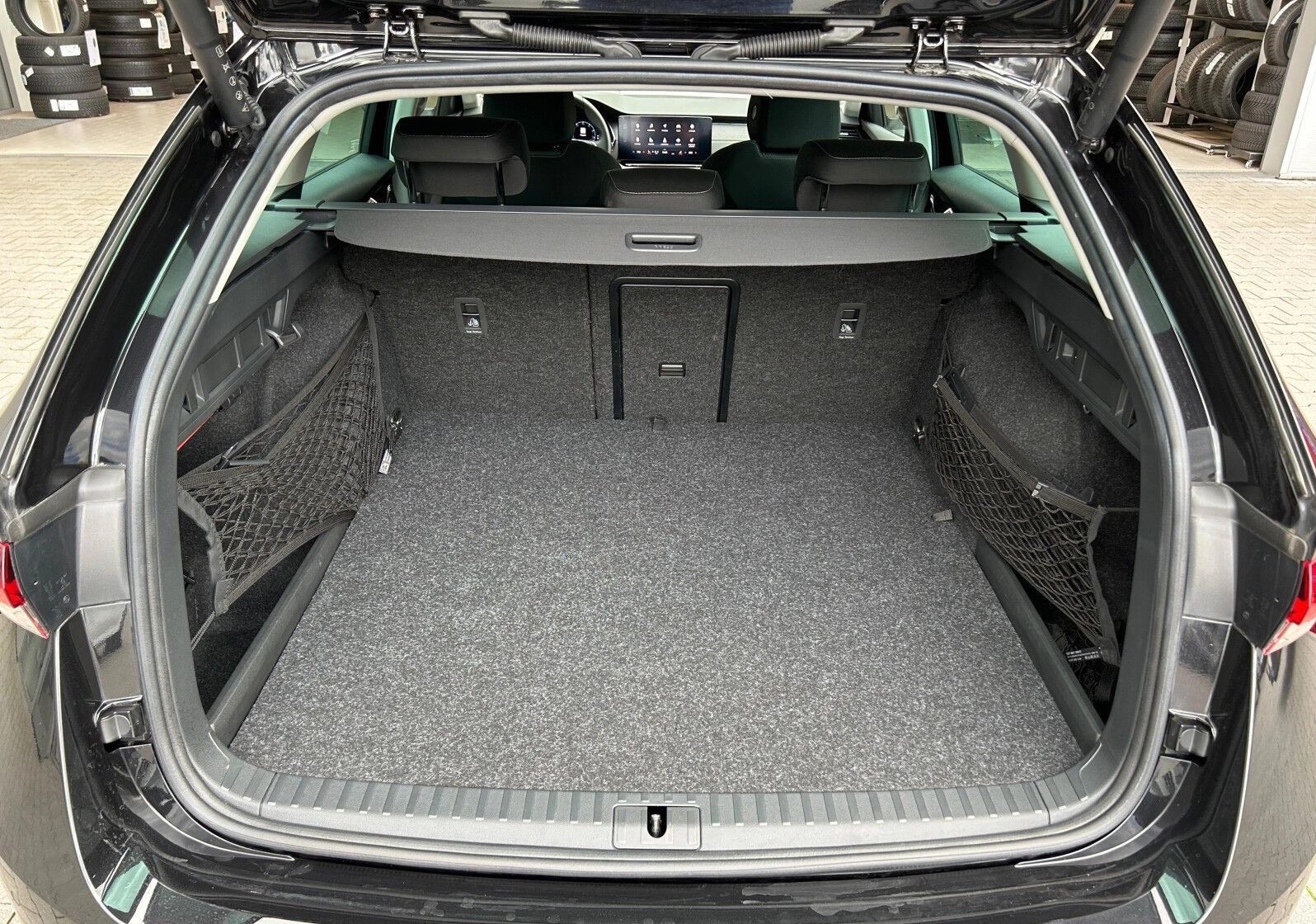 Otwarty bagażnik czarnego kombi z siatkami po bokach i szarą wykładziną. Widoczny rząd opon w tle. Przestrzeń ładunkowa gotowa do transportu bagaży.