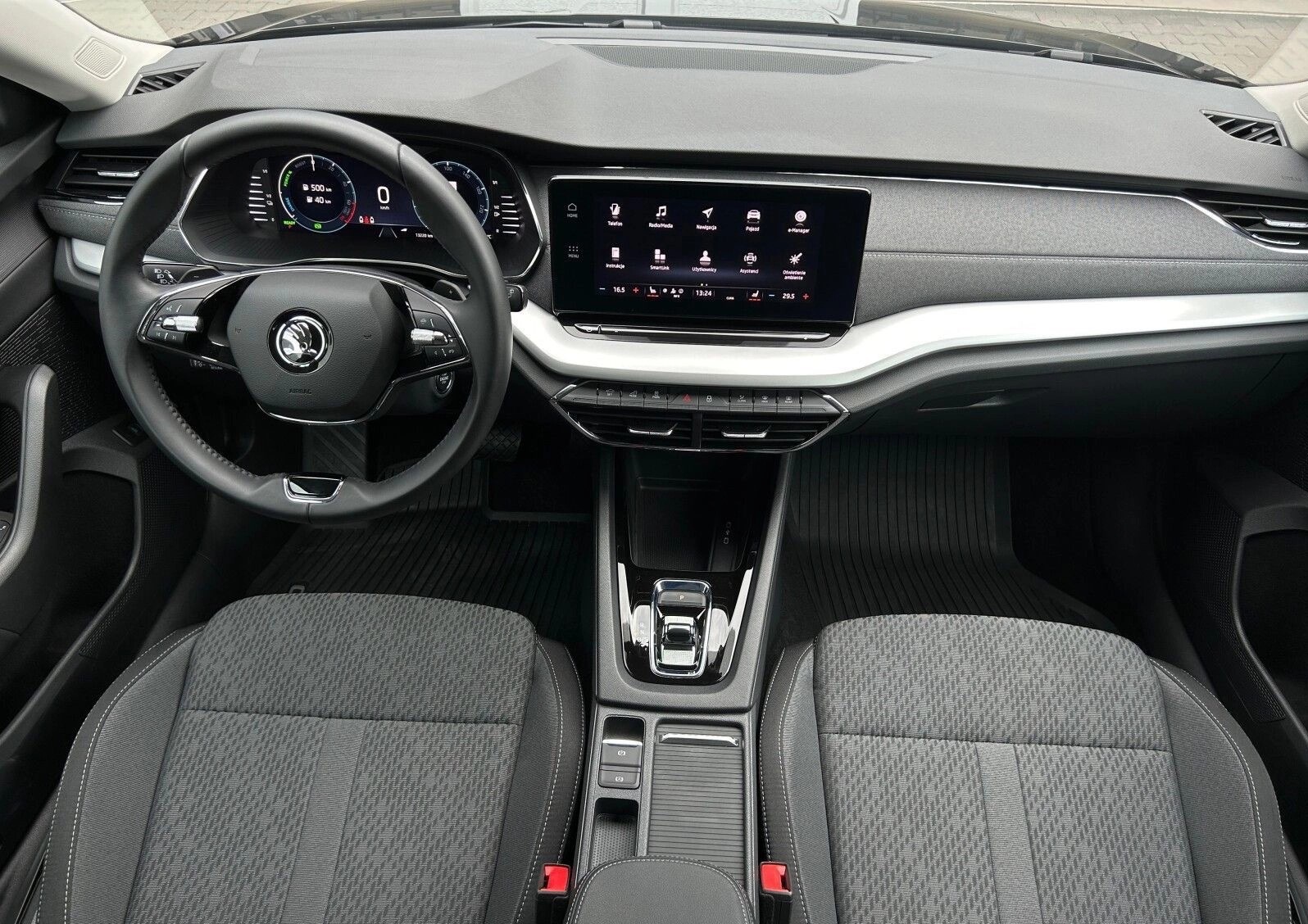Wnętrze nowoczesnego samochodu Skoda Octavia: deska rozdzielcza z dotykowym ekranem, kierownica z logo, fotele z szarej tkaniny. Widok z perspektywy pasażera.