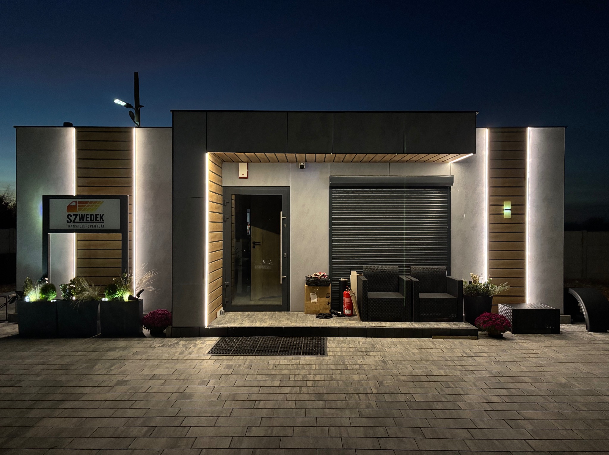 Nowoczesny budynek biurowy oświetlony ledami, z widoczną kamerą monitoringu nad wejściem. Na zewnątrz rośliny w donicach i dwa fotele. Nocne ujęcie.