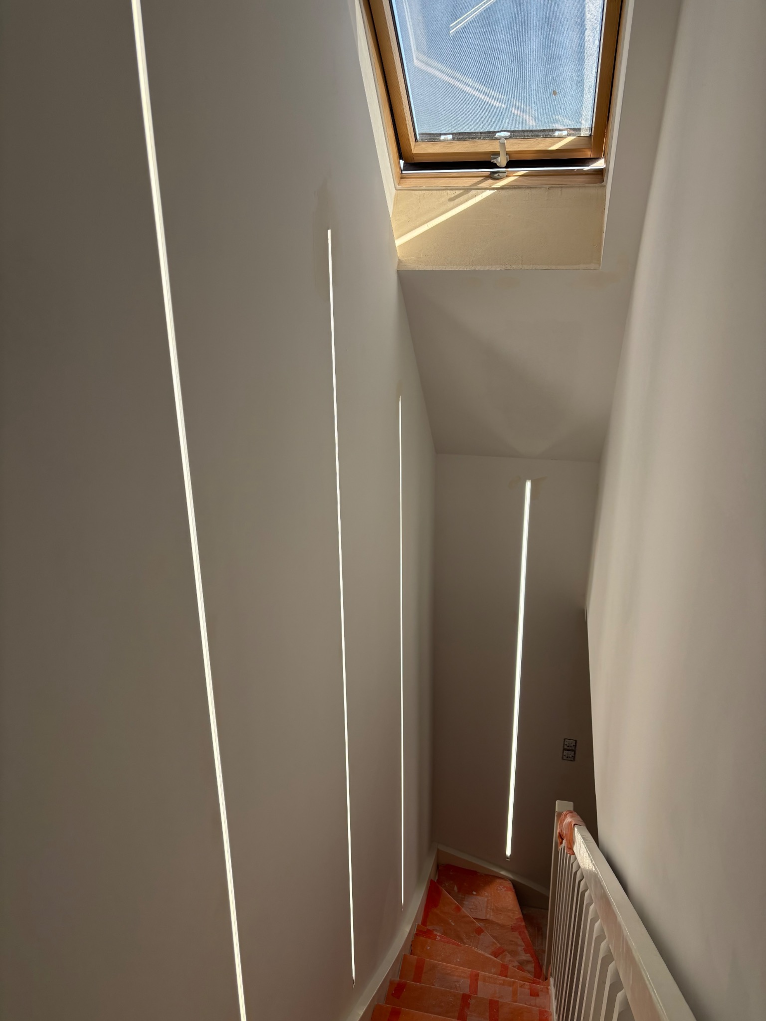 Wnętrze klatki schodowej z oknem dachowym i pionowymi, wpuszczanymi oprawami oświetleniowymi LED na ścianach. Schody zabezpieczone folią ochronną.