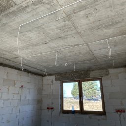 Surowy stan: białe ściany z betonu komórkowego, okno z widokiem na zewnątrz i poprowadzone kable elektryczne pod sufitem w nowo budowanym domu.