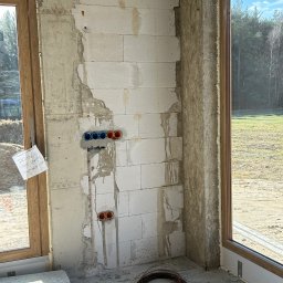 Ściana z pustaków z przygotowanymi otworami pod instalację elektryczną, widoczne kable i puszki. Okno z widokiem na zewnątrz. W tle las.