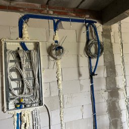 VoltBit - Rozdzielnia elektryczna z przewodami w niebieskich peszlach, zamontowana na ścianie z betonu komórkowego. Przewody zabezpieczone pianką montażową.