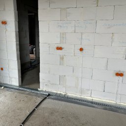 VoltBit - Instalacja elektryczna w surowym wnętrzu: pomarańczowe puszki podtynkowe na ścianie z bloczków, koryta kablowe przy podłodze, przewody. Etap prac przed wykończeniem.