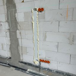 VoltBit - Wnęka w ścianie z bloków, wypełniona pianką montażową, z widocznymi pomarańczowymi puszkami instalacyjnymi i przewodami. Etap prac instalacyjnych w surowym stanie.