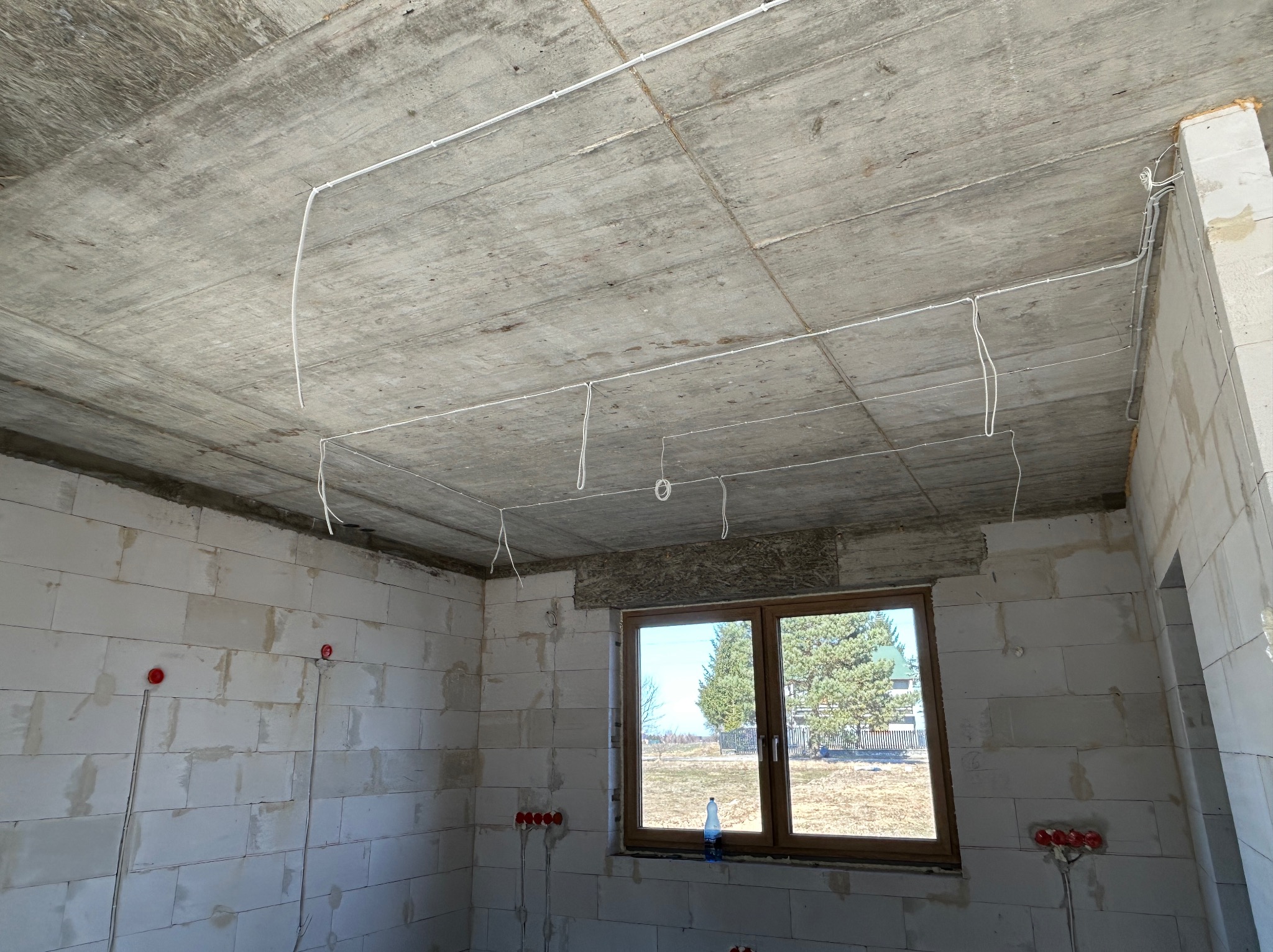 Surowy stan: białe ściany z betonu komórkowego, okno z widokiem na zewnątrz i poprowadzone kable elektryczne pod sufitem w nowo budowanym domu.