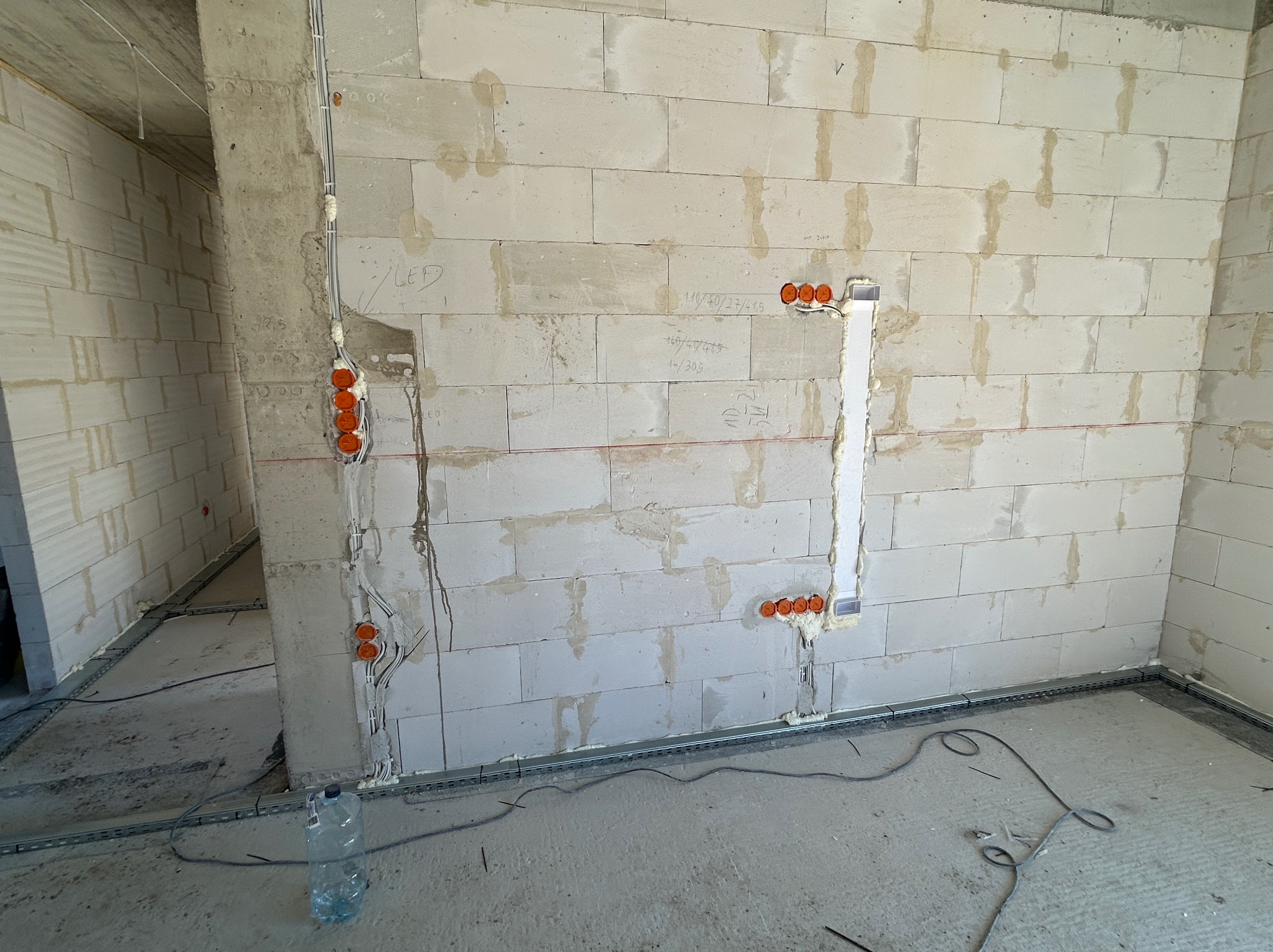 Wewnętrzna instalacja elektryczna w surowym budynku z białych bloczków. Widoczne kable w peszlach, puszki elektryczne i metalowe korytka kablowe przy podłodze.