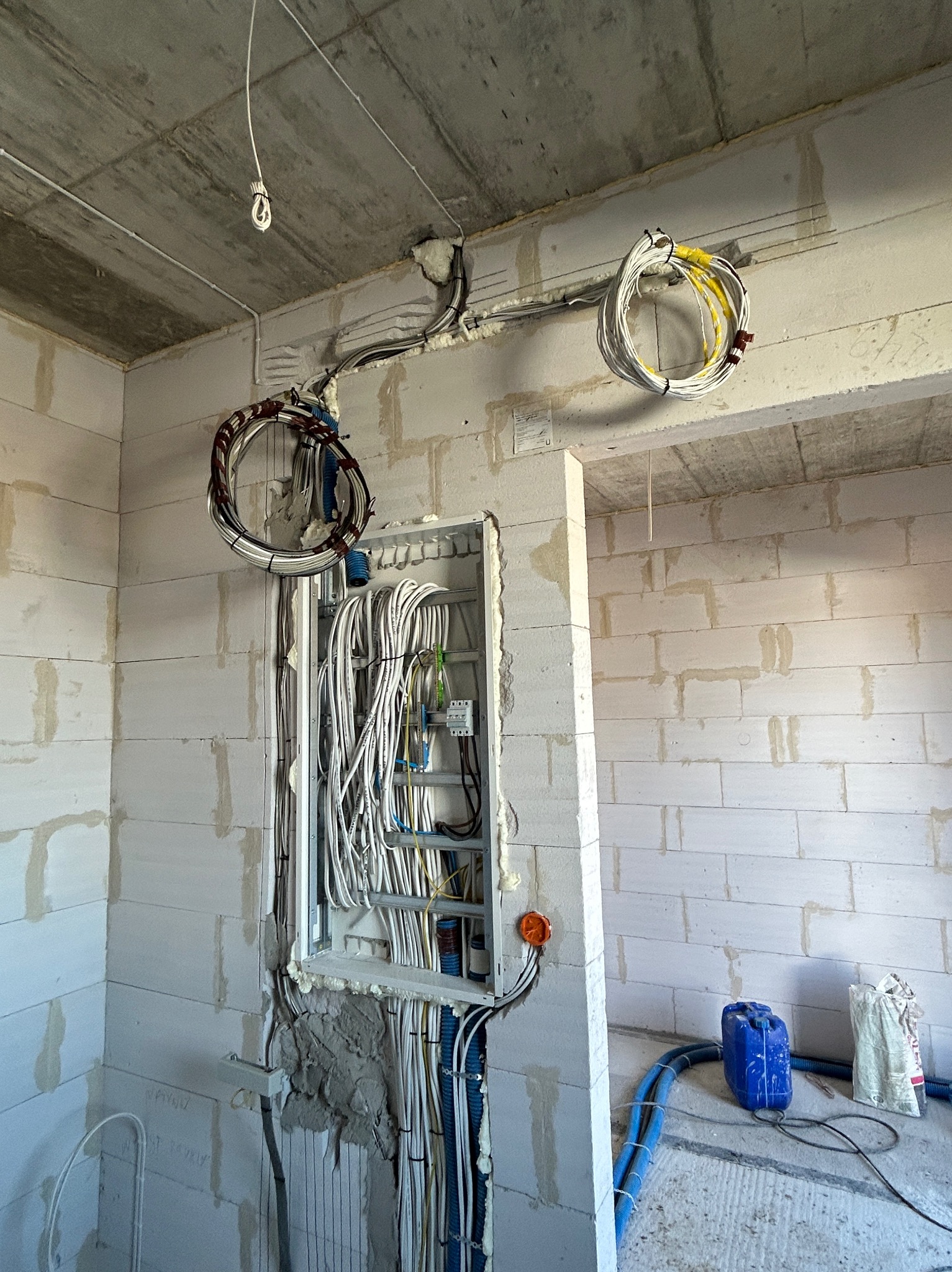 Wnętrze w trakcie prac: widoczne przewody elektryczne ułożone w ścianie z białego bloczka, zwisające kable i niebieskie rury na podłodze. Surowy stan wykończenia.