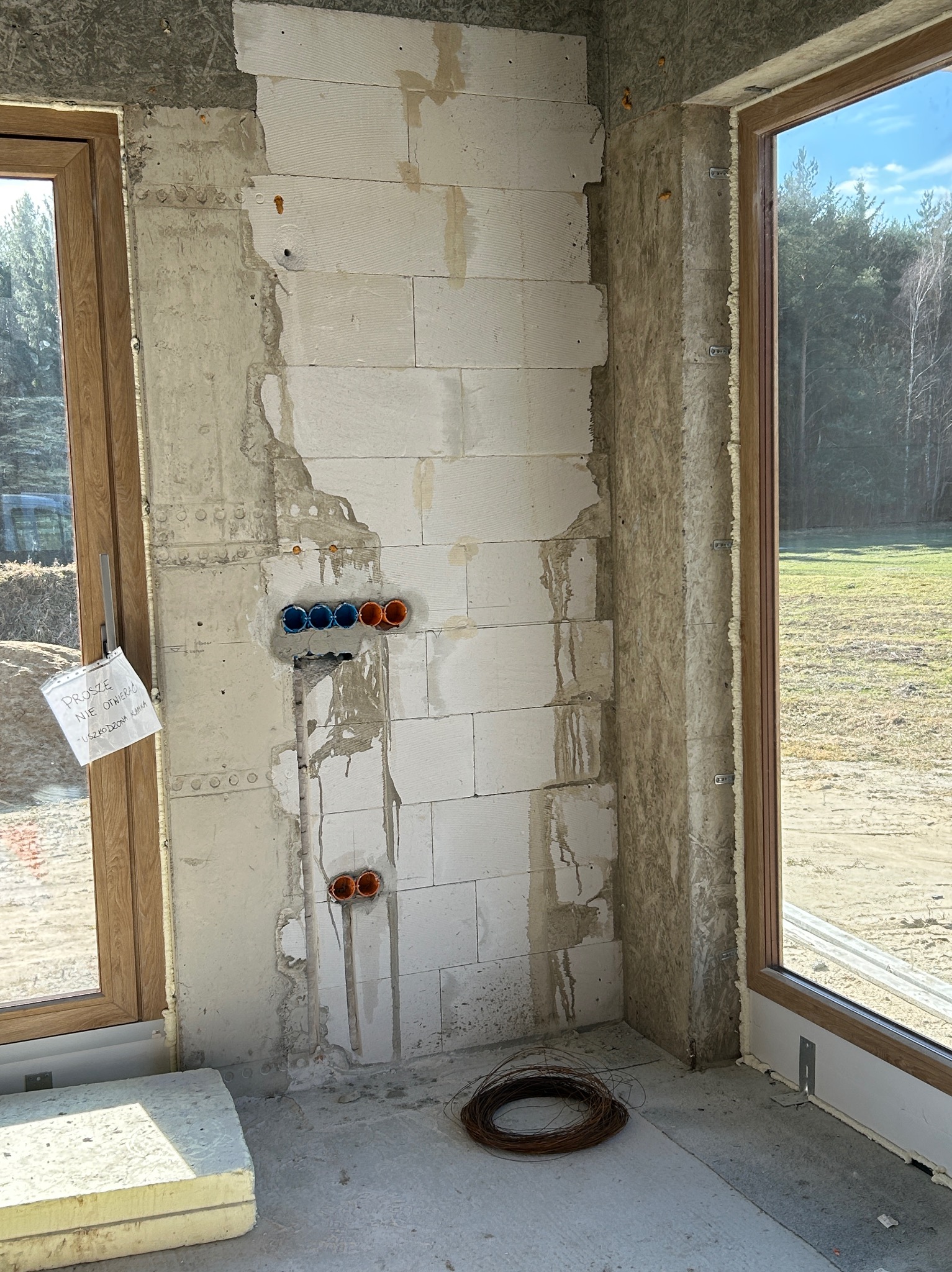 Ściana z pustaków z przygotowanymi otworami pod instalację elektryczną, widoczne kable i puszki. Okno z widokiem na zewnątrz. W tle las.