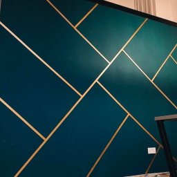 OU Kreacje - Metaliczna, ciemnozielona ściana z geometrycznym wzorem ze złotych pasków. Dekoracyjne malowanie z połyskiem, widoczny fragment podłogi i gniazdko elektryczne.