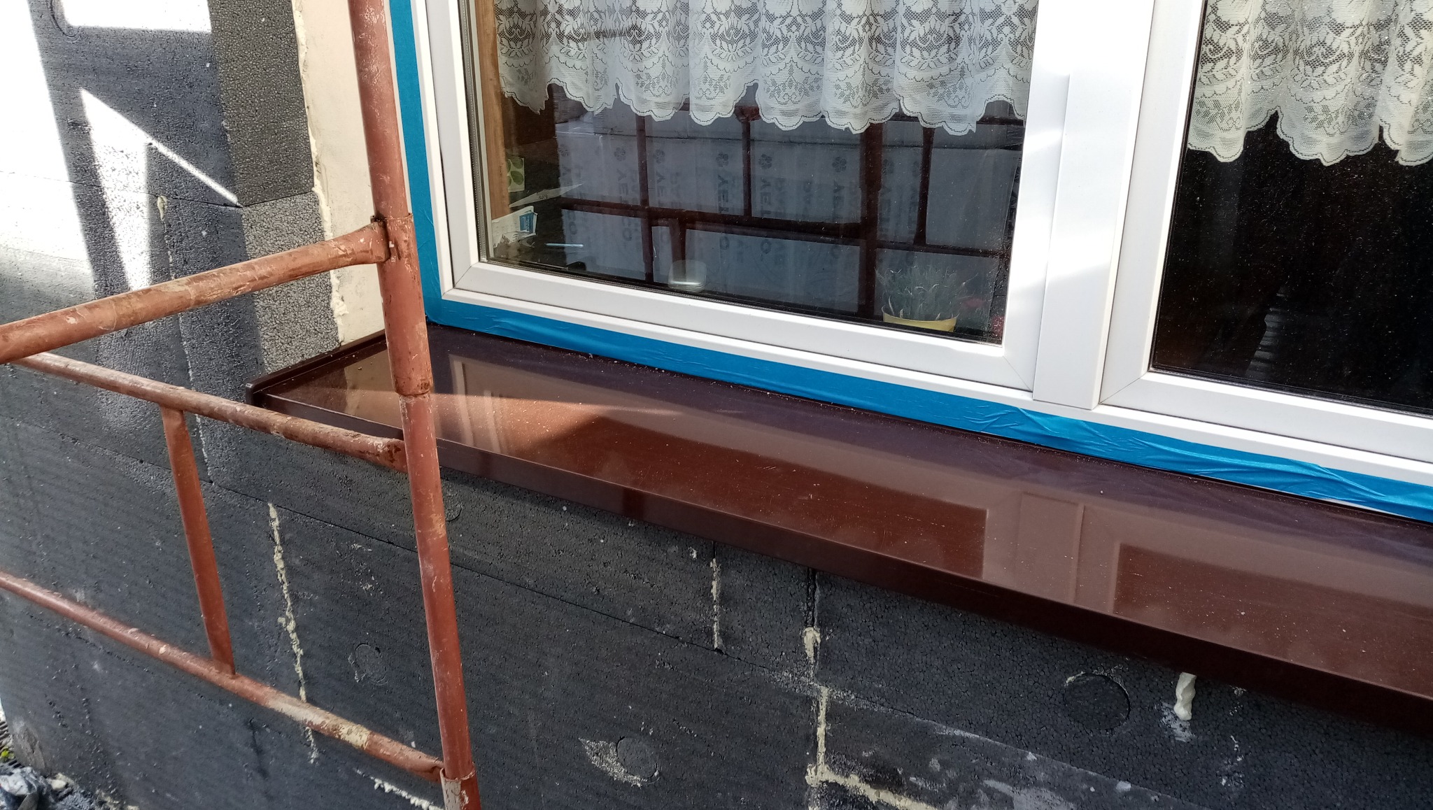 Okna z brązowym parapetem zewnętrznym, zabezpieczone folią, na tle ocieplonej ściany z widocznym rusztowaniem. Detal prac elewacyjnych.