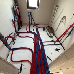 Adrian Woźny Usługi Hydrauliczne - Rozłożone rury ogrzewania podłogowego w kolorze czerwonym i niebieskim na białej izolacji, widok z góry, surowy stan wykończenia wnętrza.