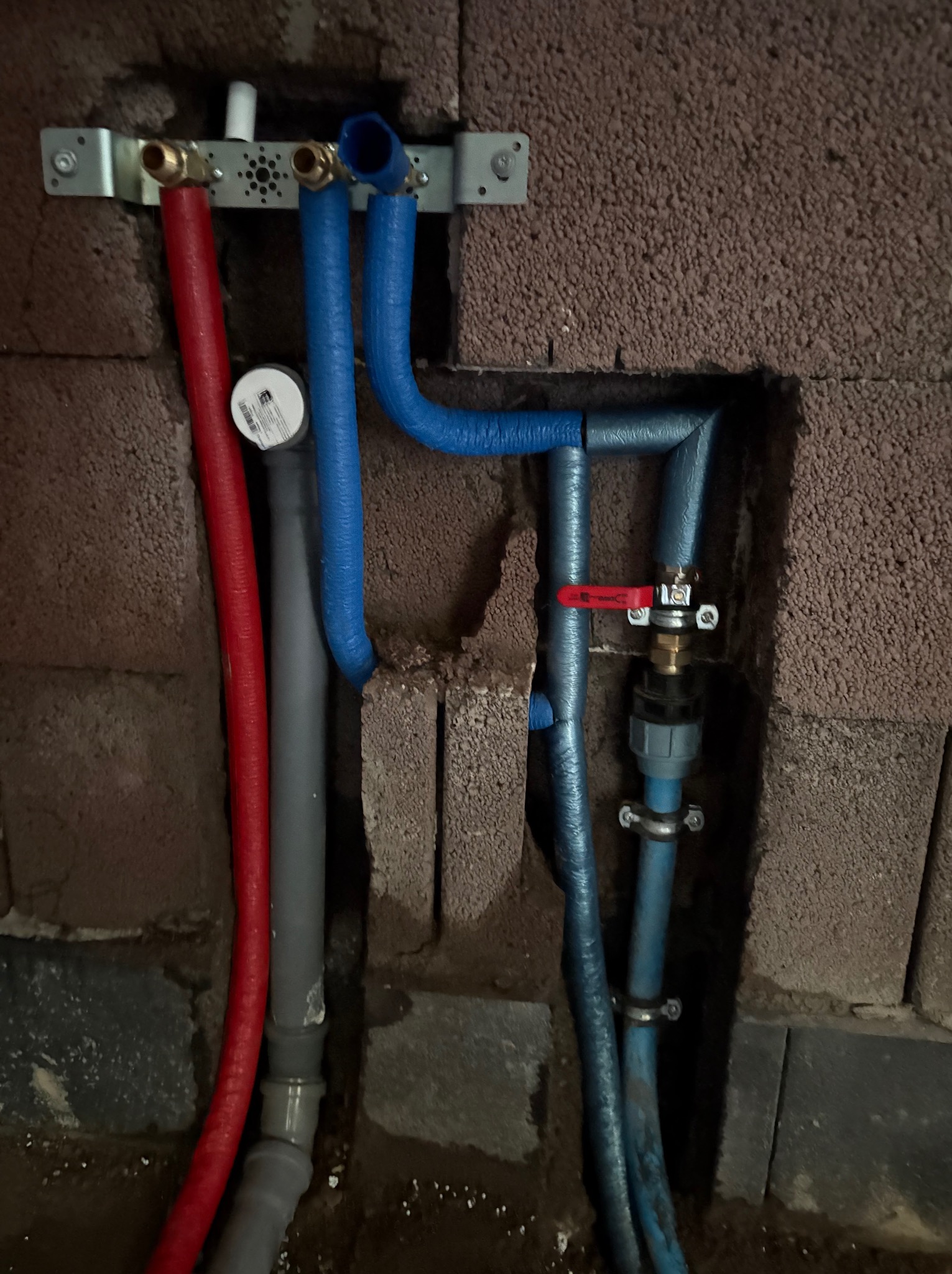 Instalacja wodna z rurami w kolorze czerwonym i niebieskim, zamontowana na ścianie z cegieł. Widoczny zawór z czerwoną rączką i metalowy uchwyt mocujący.