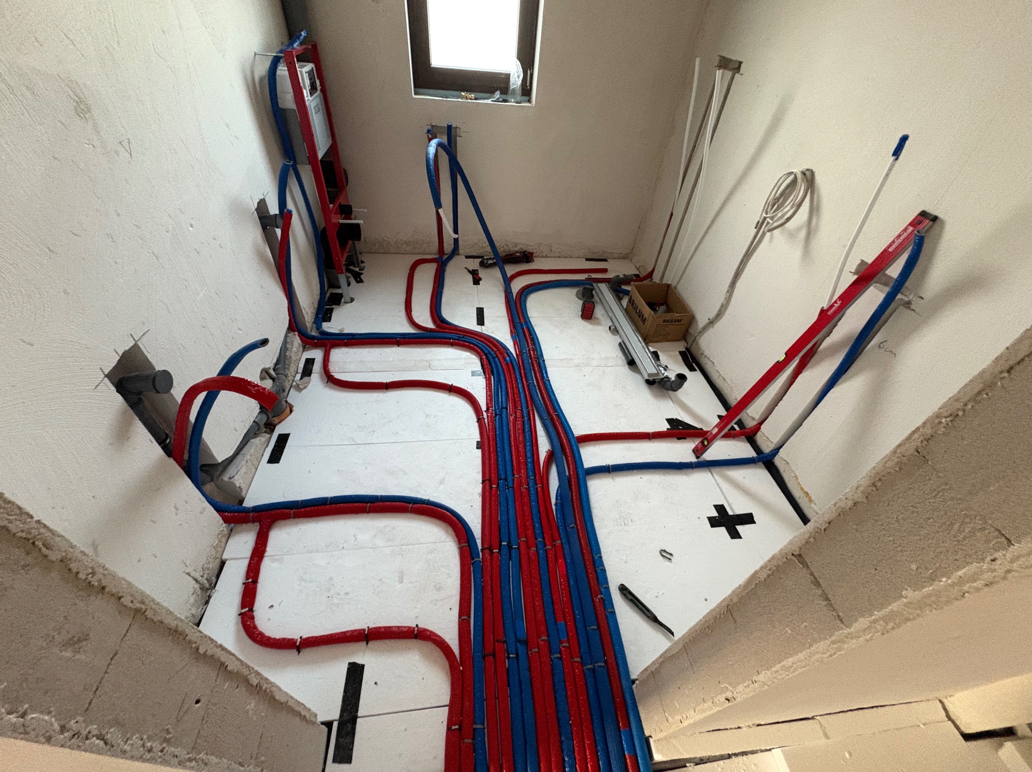 Rozłożone rury ogrzewania podłogowego w kolorze czerwonym i niebieskim na białej izolacji, widok z góry, surowy stan wykończenia wnętrza.