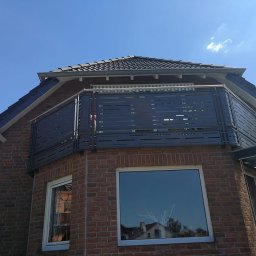 Natis Paweł Stankiewicz - Szara, nowoczesna balustrada balkonowa z poziomymi szczelinami, zamontowana na ceglanym budynku z widokiem na błękitne niebo. Detal architektoniczny.