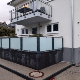 Natis Paweł Stankiewicz - Nowoczesna balustrada balkonowa z matowego szkła i metalu, zamontowana na niskiej, ciemnej podmurówce. Widok z zewnątrz, na tle jasnej elewacji budynku.