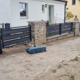 Natis Paweł Stankiewicz - Montaż nowoczesnego ogrodzenia panelowego z elementami kamiennymi. Widoczne narzędzia i materiały budowlane. Dwóch pracowników w trakcie pracy.