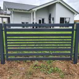 Natis Paweł Stankiewicz - Nowoczesne, poziome ogrodzenie panelowe w kolorze grafitowym, na tle domu z szarą elewacją i zielonym trawnikiem. Minimalistyczny design, świeżo posadzona trawa.