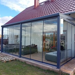 Natis Paweł Stankiewicz - Szklana zabudowa balkonu z przesuwnymi drzwiami, widok z zewnątrz. Wnętrze z meblami wypoczynkowymi. Czerwona dachówka domu w tle, błękitne niebo.