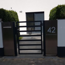 Natis Paweł Stankiewicz - Nowoczesna, czarna furtka z numerem domu '42 Schleusenweg', wkomponowana w ogrodzenie z tujami. Minimalistyczny design, jasna elewacja budynku w tle.