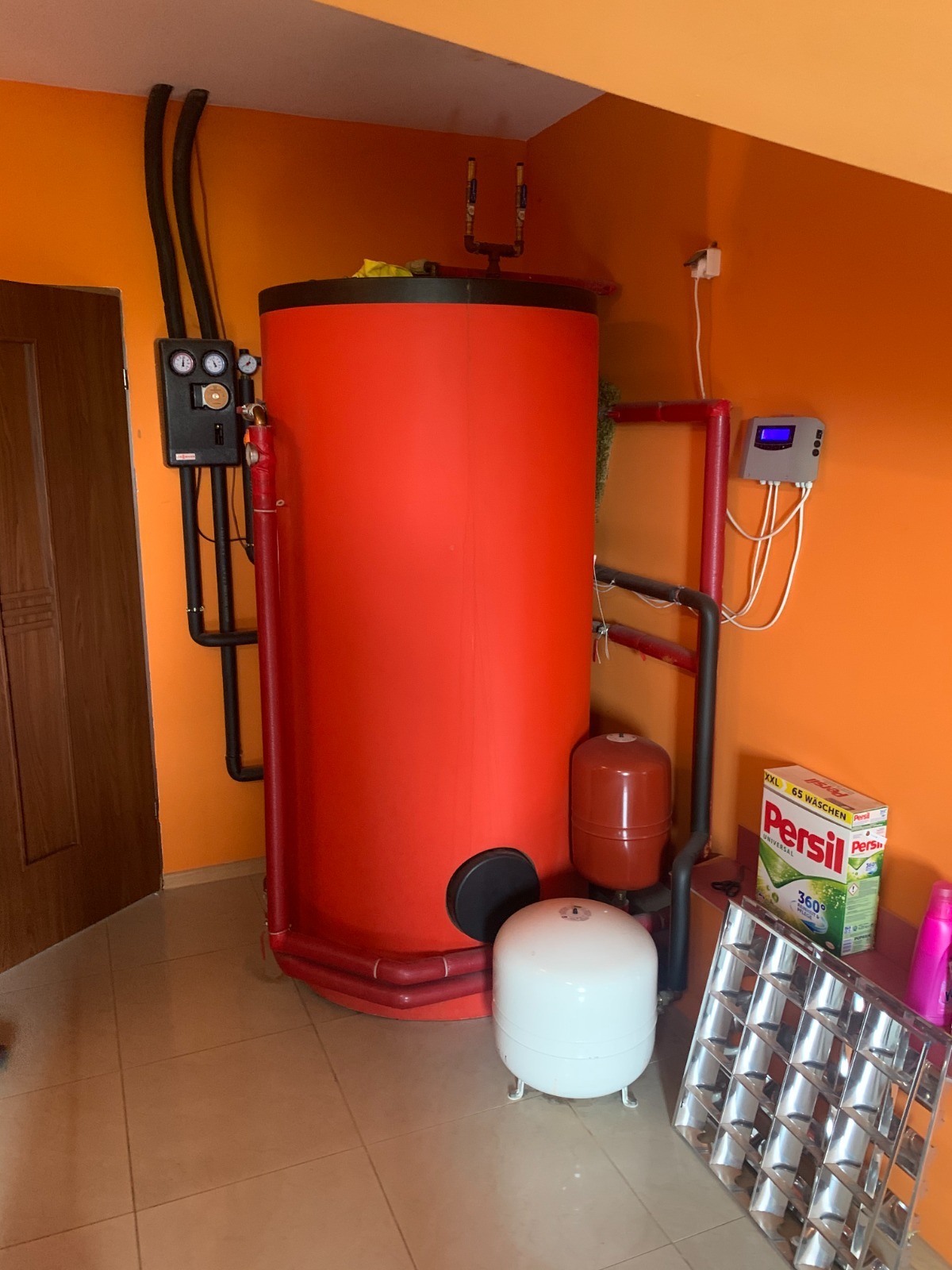 Czerwony, cylindryczny zbiornik ciepłej wody użytkowej z czarnymi rurami i osprzętem na tle pomarańczowej ściany. Widoczne elementy instalacji i akcesoria.