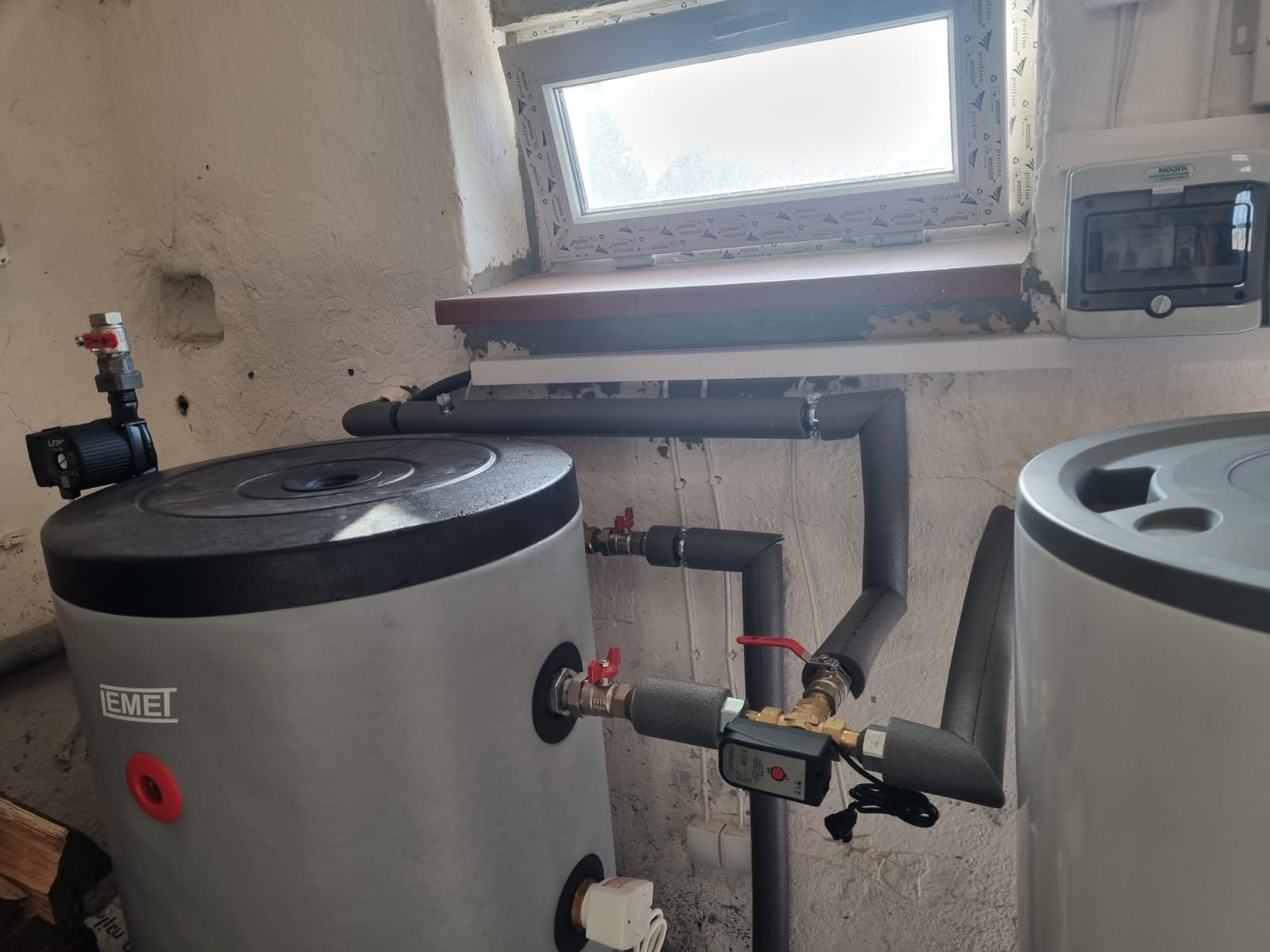 Szary zbiornik akumulacyjny Emet z izolowanymi rurami i zaworami w kotłowni, widoczne okno i skrzynka elektryczna. Ujęcie z bliska, skupione na instalacji.