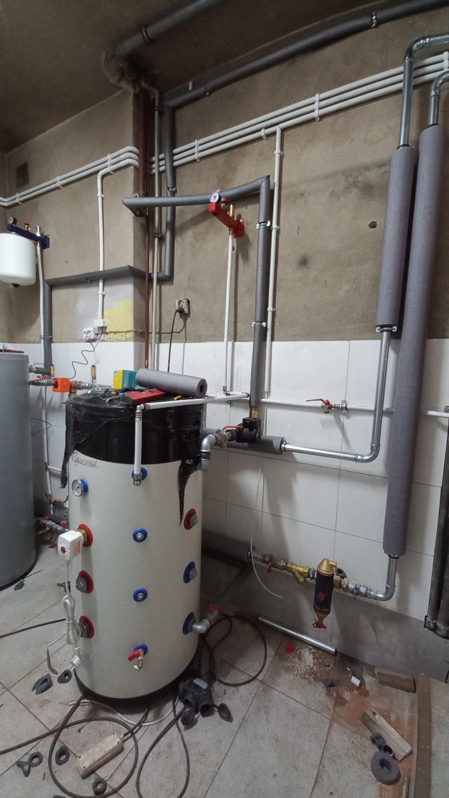 Bojler z podłączoną instalacją hydrauliczną i grzewczą w pomieszczeniu z wykończeniem w stylu retro. Widoczne rury, zawory i izolacja termiczna.