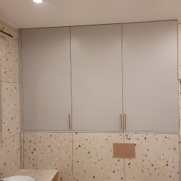 Interier - Szare szafki łazienkowe na wymiar z złotymi uchwytami, dopasowane do ściany z wzorem lastryko. Umywalka z blatem i fragment toalety w kadrze.