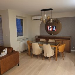 Interier - Jasne wnętrze jadalni z drewnianym stołem i krzesłami, nad którymi wisi designerska lampa. W tle szafka i okrągłe lustro z telewizorem.