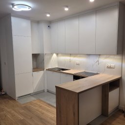 Interier - Nowoczesna kuchnia na wymiar z białymi szafkami i drewnianym blatem, oświetlona punktowo, z marmurowym panelem nad blatem i wyspą kuchenną.