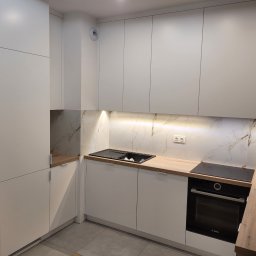 Interier - Nowoczesna, jasna kuchnia z białymi frontami i drewnianym blatem. Marmurowe wykończenie ściany nad blatem. Widoczny zlew, płyta indukcyjna i piekarnik.