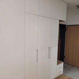 Interier - Biała szafa wnękowa z czterema drzwiami i górnymi szafkami, zintegrowana z siedziskiem i szufladami, w jasnym przedpokoju z drewnianą podłogą i drzwiami.