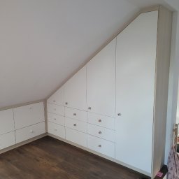 Interier - Biała szafa wnękowa na poddaszu ze skośnym sufitem, z drewnianą podłogą i dziecięcymi sandałkami. Szafki z okrągłymi uchwytami.